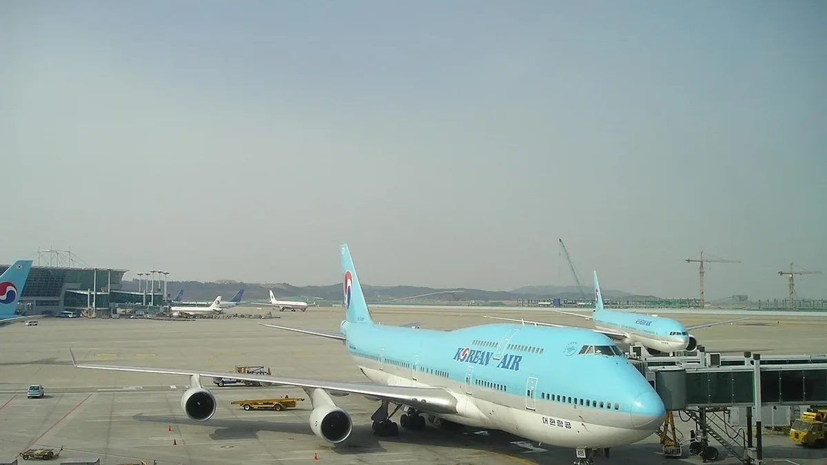A Korean Air plane at an airport, with a jetway attached to the front of the plane and a smaller plane visible in the background.