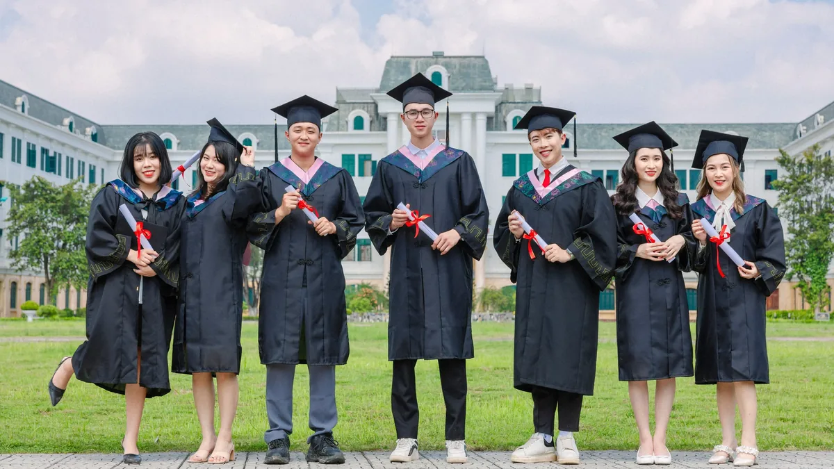 Seven young adults in graduation attire stand in a line, each holding a rolled-up diploma with a red ribbon tied around it. They are dressed